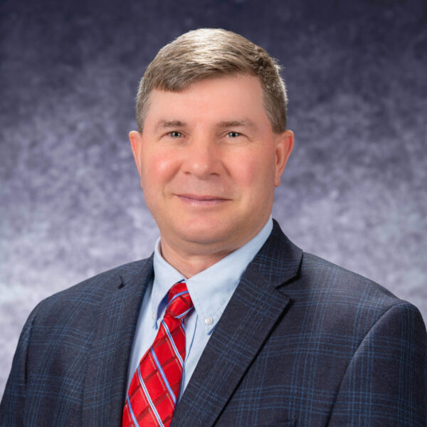 Photo of Bill Even, navy suit jacket with red checkered tie and a gray backgrouns