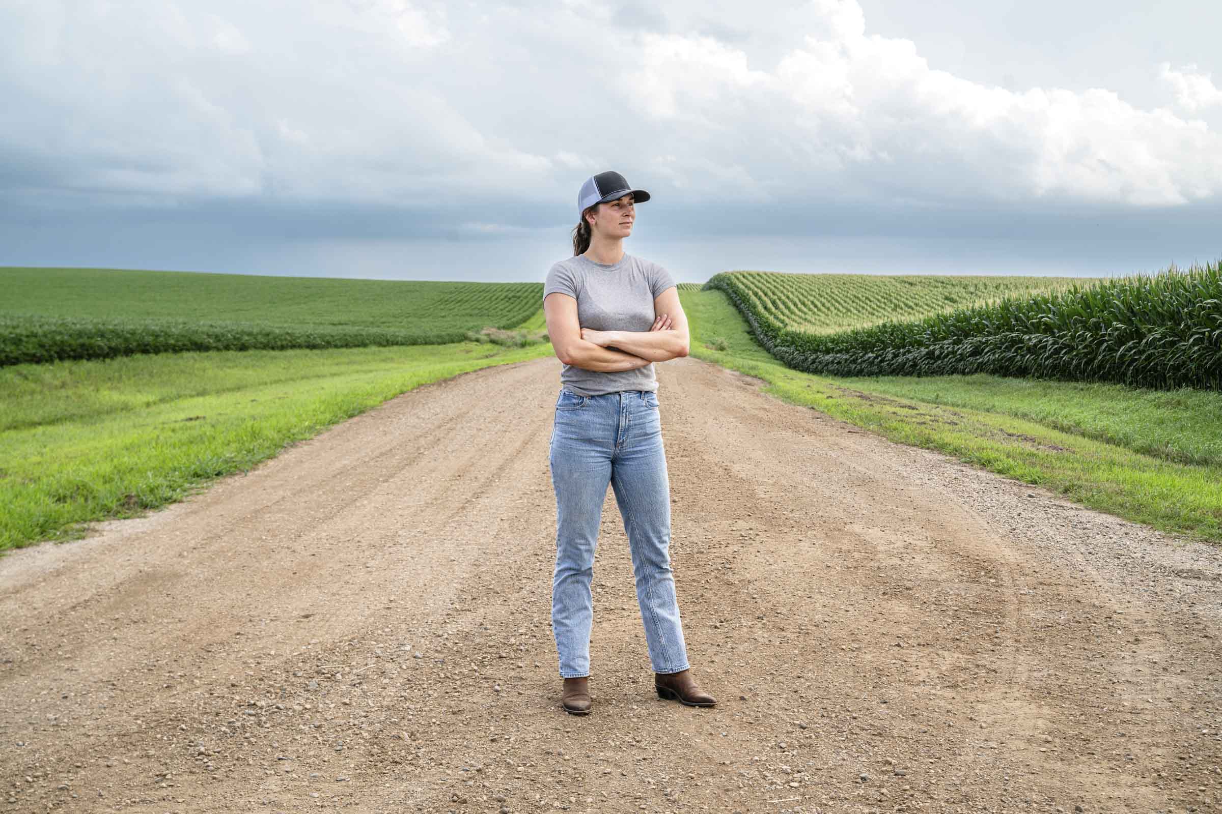 Woman standing in a dirt road, looking out over a cornfield.