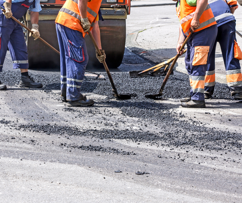 Road construction workers spreading asphalt by hand while a steamroller follows behind.