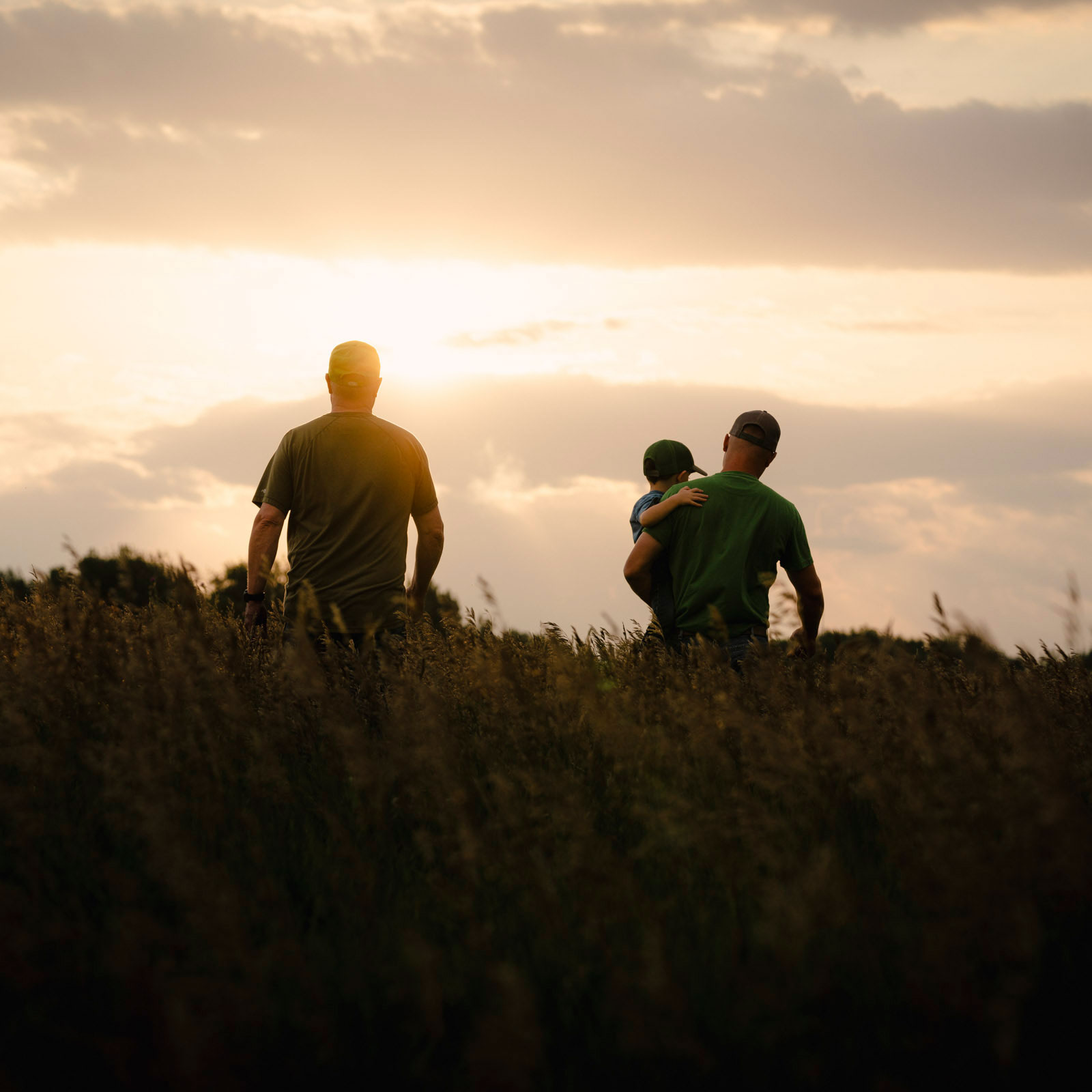 Two men walking towards sunset in a field, one is holding a child.