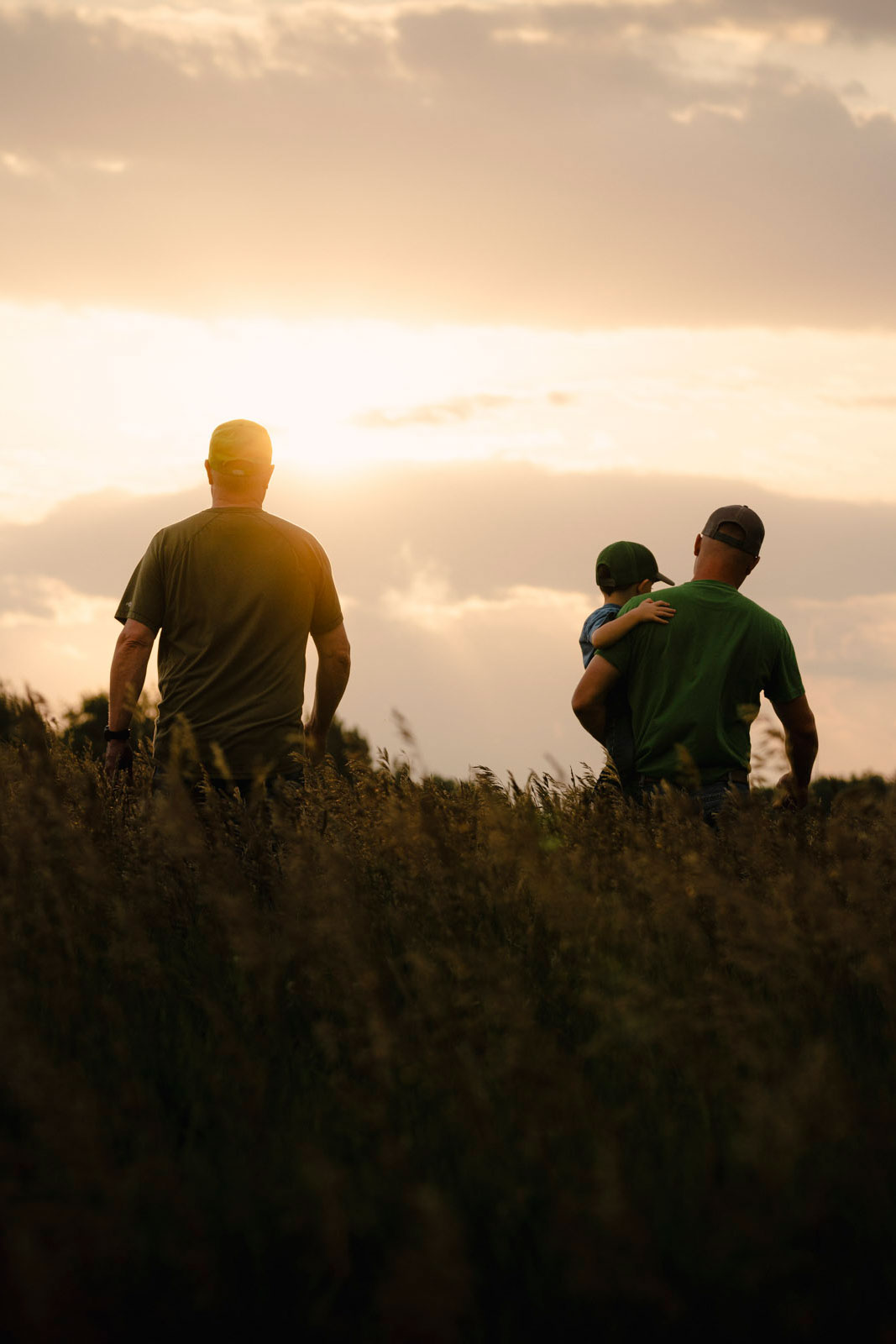 Two men walking towards sunset in a field, one is holding a child.