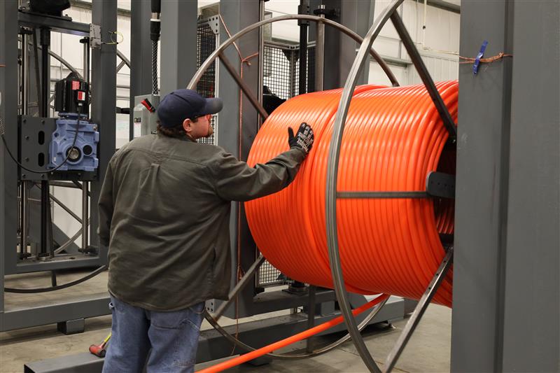 Worker guiding orange fiber conduit onto a large industrial spool during broadband infrastructure installation.