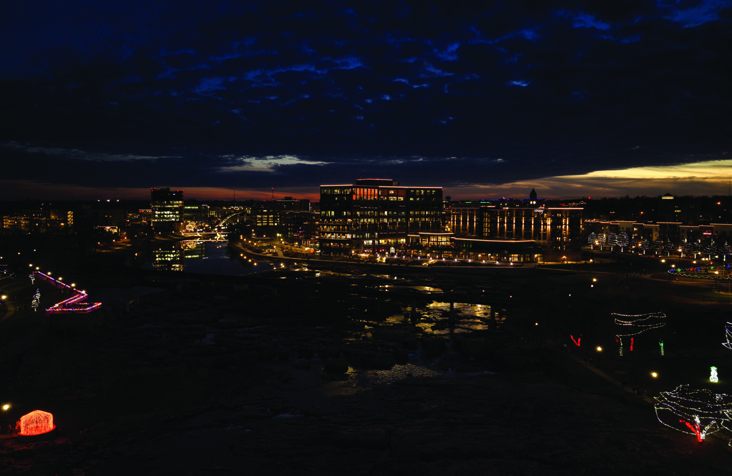 Downtown Sioux Falls skyline at dusk with Falls Park lights and illuminated buildings along the Big Sioux River