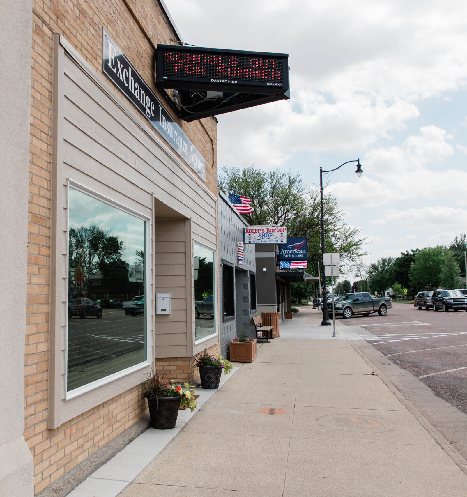 Sidewalk view of downtown Lennox, South Dakota, featuring local storefronts, business signs, and parked vehicles along Main Street.