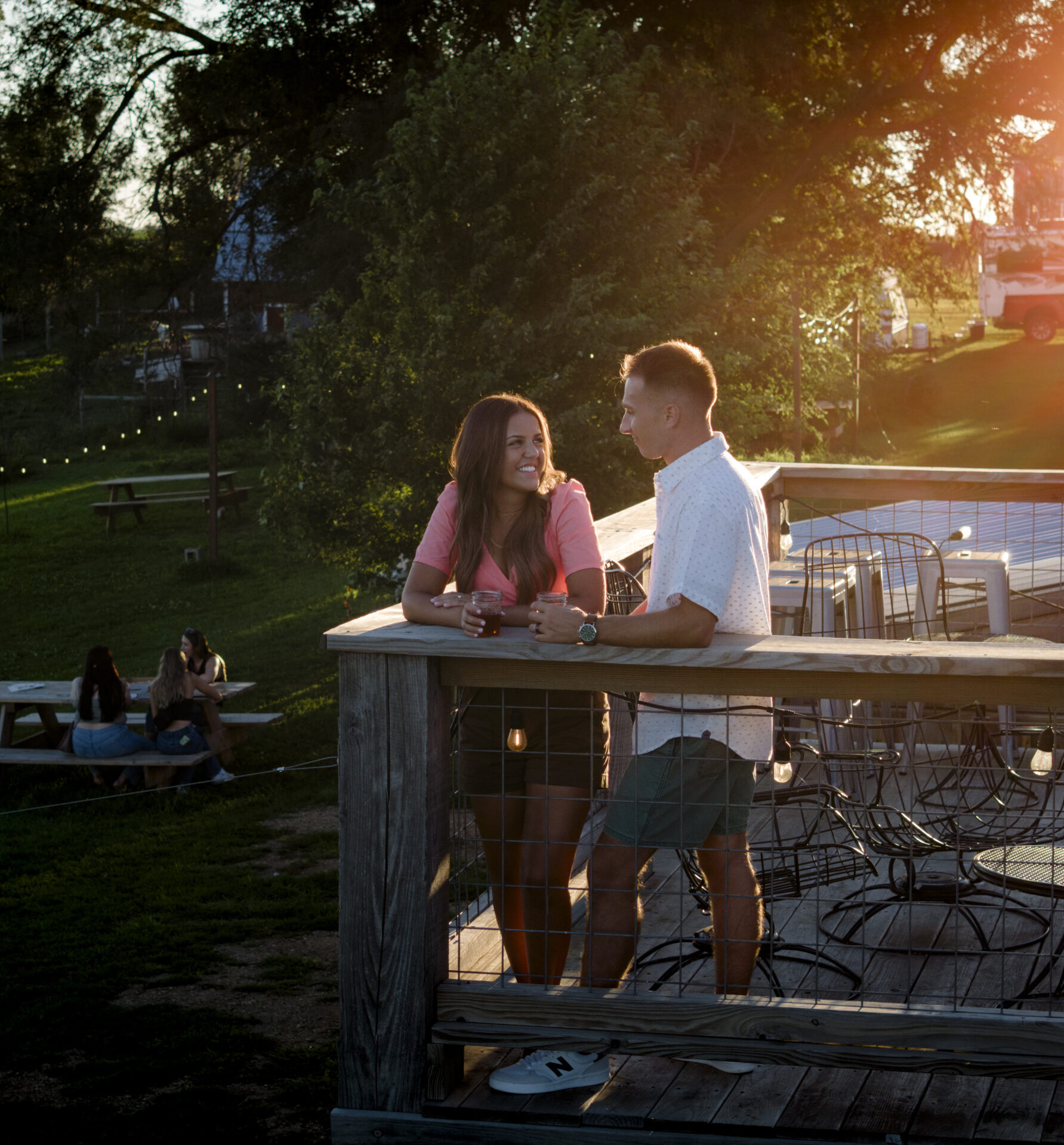 Two people stand on an outdoor deck at Good Earth Farm near Lennox, South Dakota, enjoying drinks during sunset with farmland and string lights in the background.