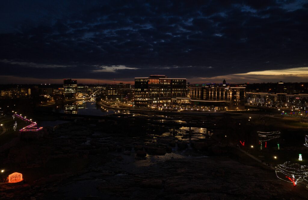 Nighttime aerial view of downtown Sioux Falls with illuminated buildings, riverfront development, and holiday lights reflecting along the Big Sioux River.