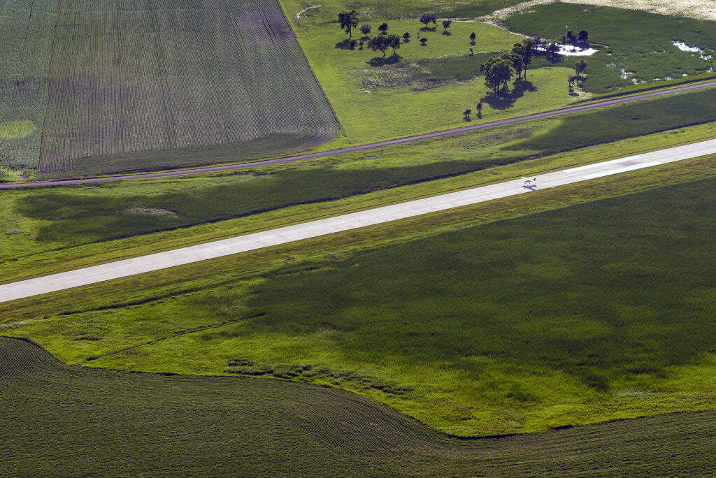 Aerial view of a small aircraft traveling along a runway surrounded by green agricultural fields, wetlands, and rural South Dakota landscape.